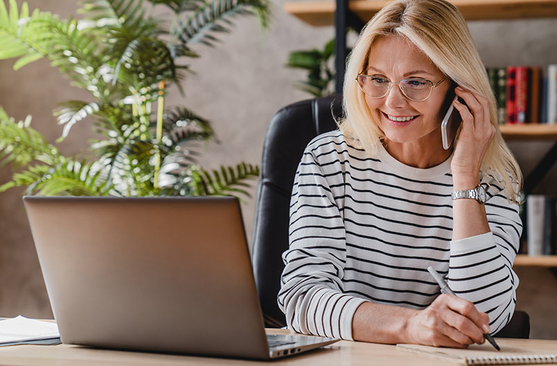 Beautiful elderly businesswoman speaking on smart phone while working and writing notes in notepad